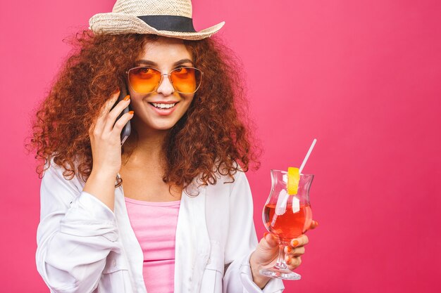 Retrato de mulher feliz em óculos de sol usando smartphone e segurando coquetel isolado em fundo rosa