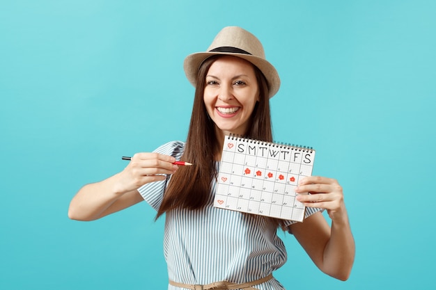 Retrato de mulher feliz em um vestido azul, chapéu segurando um lápis vermelho, calendário de períodos femininos para verificar os dias de menstruação isolados sobre fundo azul. Saúde médica, conceito ginecológico. Copie o espaço