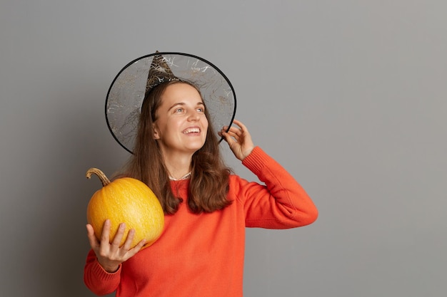 Retrato de mulher festiva encantada sorridente vestida com chapéu de bruxa segurando abóbora laranja e desviar o olhar com expressão positiva sonhando feliz Halloween posando isolado em fundo cinza