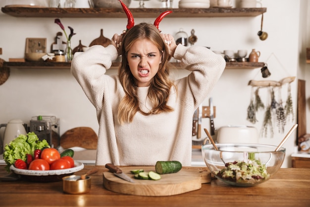 retrato de mulher fofa com raiva usando roupas casuais fazendo chifres com pimentões enquanto cozinha o jantar em uma cozinha aconchegante