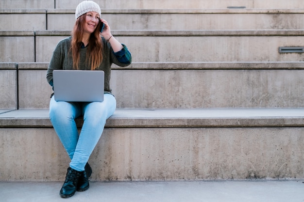 Retrato de mulher freelancer usando laptop e smartphone enquanto está sentado na escada na rua da cidade