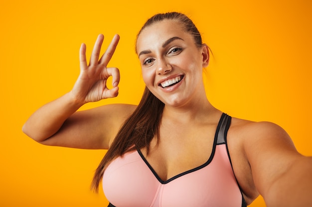 Retrato de mulher gordinha alegre em sutiã esportivo usando telefone celular para foto de selfie, isolado sobre fundo amarelo