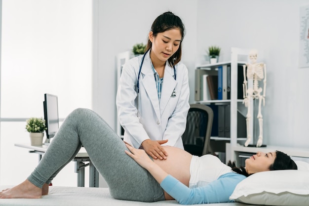 retrato de mulher grávida asiática deitada no sofá de tratamento com joelhos dobrados está recebendo fisioterapia de seu ginecologista em uma sala de clínica moderna e brilhante.