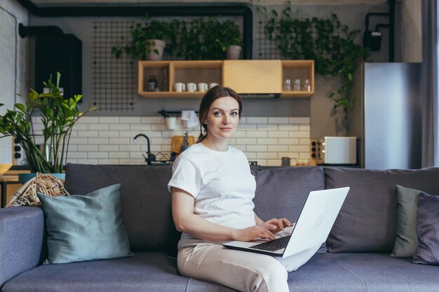 Retrato de mulher grávida em casa trabalhando remotamente com laptop online olhando para a câmera e sorrindo