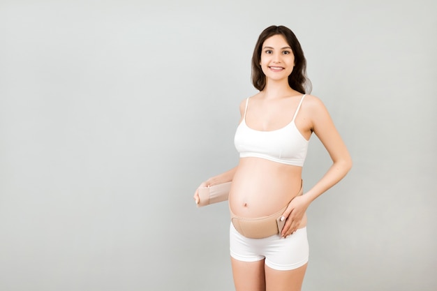 Retrato de mulher grávida em cueca vestindo faixa elástica de maternidade no fundo cinza com espaço de cópia. Conceito de cinto de suporte abdominal ortopédico.