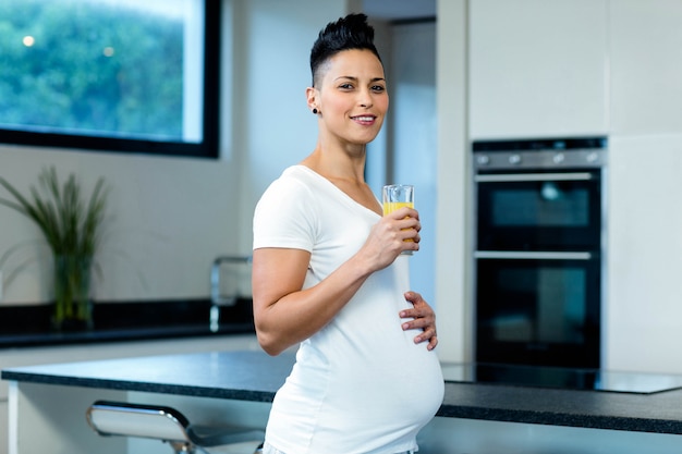 Retrato de mulher grávida em pé com a mão na barriga e bebendo suco na cozinha