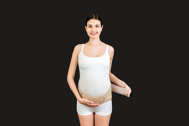 Retrato de mulher grávida vestindo curativo ortopédico para reduzir a dor nas costas em fundo preto com espaço de cópia. Conceito de cinto de suporte abdominal ortopédico.