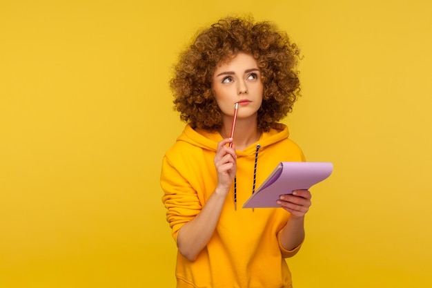 Retrato de mulher hipster de cabelos encaracolados pensativa com capuz estilo urbano pensando sobre a ideia inteligente, segurando o lápis e o caderno para escrever planos, lista de tarefas. tiro de estúdio interno isolado em fundo amarelo