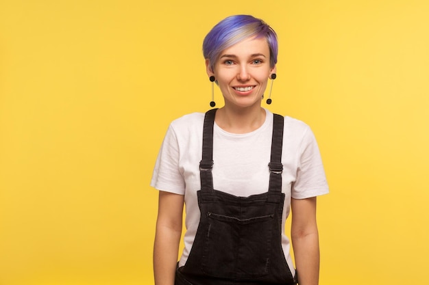 Retrato de mulher hipster na moda feliz com cabelo curto violeta de macacão olhando para a câmera com sorriso positivo humor elegante elegante aparência isolada em fundo amarelo studio shot