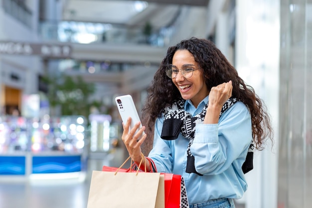 Retrato de mulher hispânica feliz compradora dentro do shopping usa smartphone navega por descontos on-line