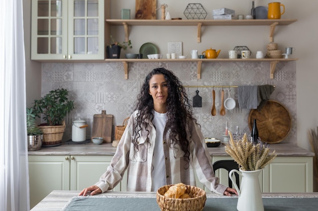 Retrato de mulher hispânica feliz em casa na cozinha mulher com cabelo encaracolado sorrindo e olhando para