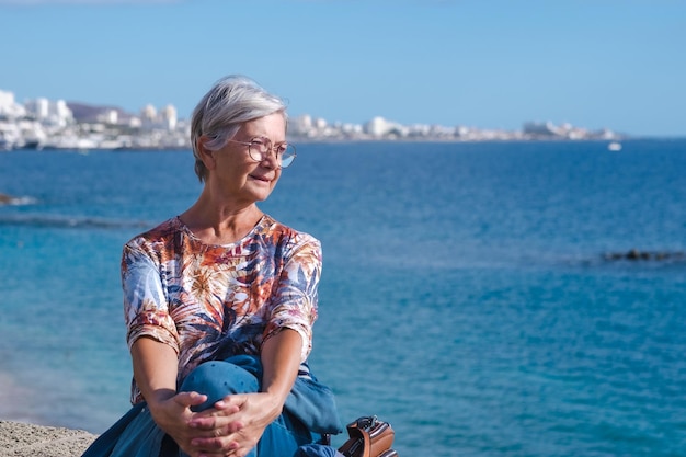 Retrato de mulher idosa atraente sentada na praia olhando para longe no horizonte sobre o mar Mulher caucasiana sênior relaxada aproveitando férias ou aposentadoria