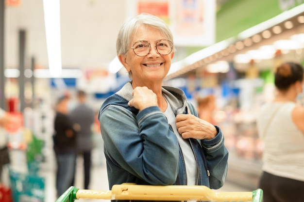 Retrato de mulher idosa sorridente fazendo compras no supermercado com carrinho esperando sua vez de carne e presunto cliente idoso caucasiano em uma mercearia