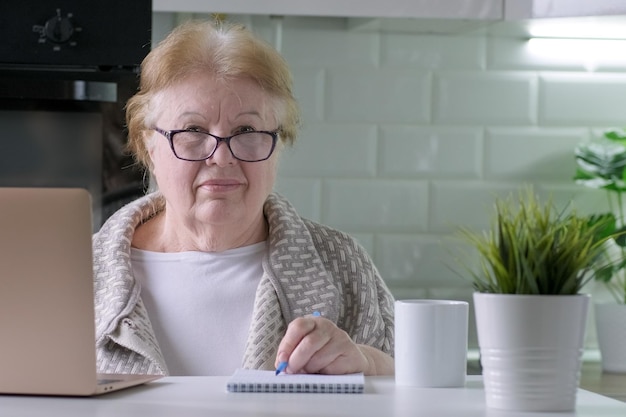 Retrato de mulher idosa sorridente usando óculos sentado na mesa com o laptop em casa