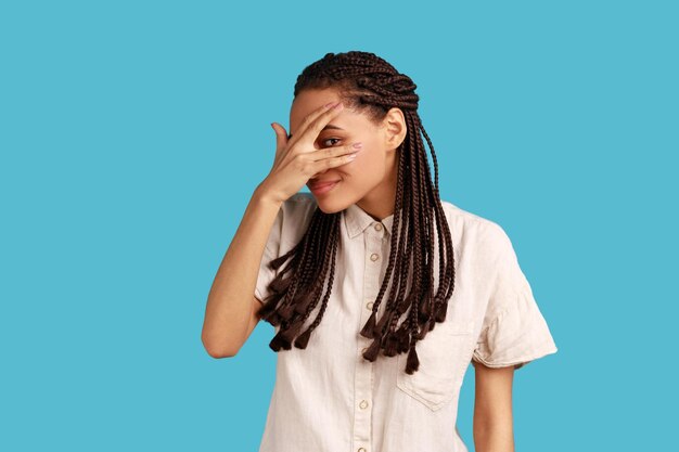 Foto retrato de mulher interessada com dreadlocks cobrindo os olhos com a mão espreitando por entre os dedos com expressão curiosa feliz vestindo camisa branca foto de estúdio interior isolada em fundo azul