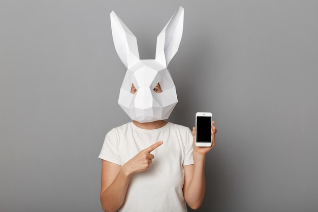 Retrato de mulher irreconhecível vestindo camiseta branca e máscara de coelho de papel em pé isolado sobre fundo cinza mostrando telefone inteligente com tela vazia apontando para o espaço da cópia