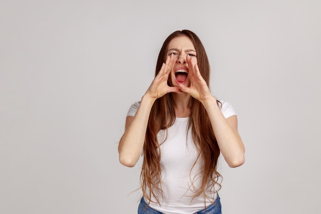 Retrato de mulher irritada com cabelo escuro em pé e gritando sobre más notícias ou problemas segurando as mãos perto da boca vestindo camiseta branca Foto de estúdio interior isolada em fundo cinza