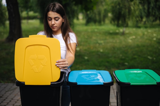 Foto retrato de mulher jogando uma garrafa de água de plástico vazia em uma lixeira, uma árvore, lixeiras para reciclagem ao ar livre