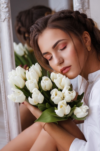 Retrato de mulher jovem bonita no vestido branco, olhando seu reflexo no espelho e segurando flores da primavera. linda garota natural posando contra espelho com buquê de tulipas. dia das mulheres