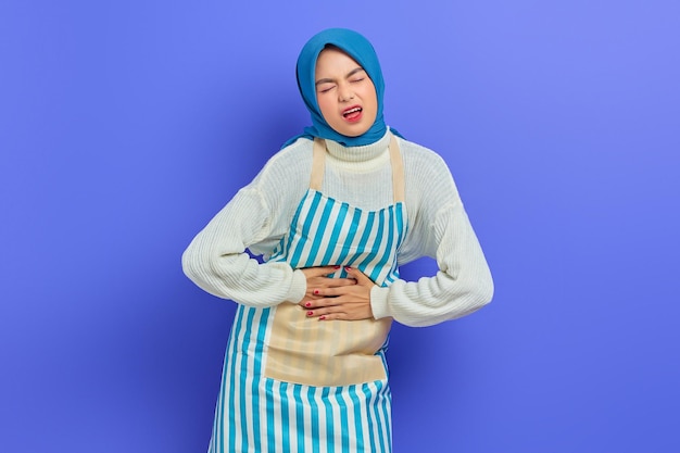 Retrato de mulher jovem dona de casa doente em hijab e avental listrado Tendo dor abdominal colocar as mãos no estômago no fundo roxo Pessoas dona de casa conceito de estilo de vida muçulmano