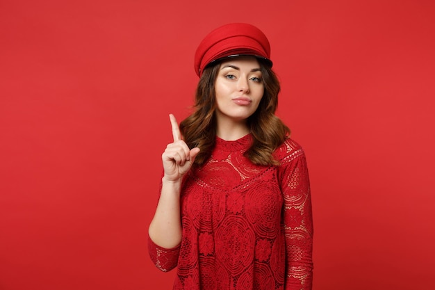 Retrato de mulher jovem e atraente no vestido de renda e boné apontando o dedo indicador para cima olhando a câmera isolada no fundo da parede vermelha brilhante. Emoções sinceras de pessoas, conceito de estilo de vida. Mock up espaço de cópia.