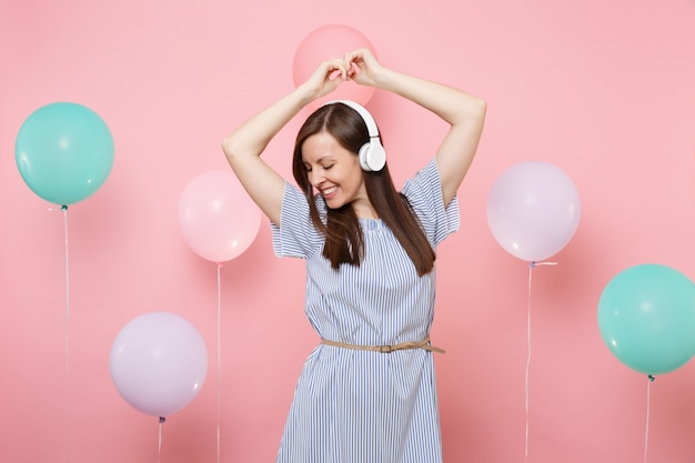 Retrato de mulher jovem e atraente sorridente com fones de ouvido no vestido azul, ouvindo música dançando no fundo rosa pastel com balões de ar coloridos. Festa de aniversário, emoções sinceras de pessoas.