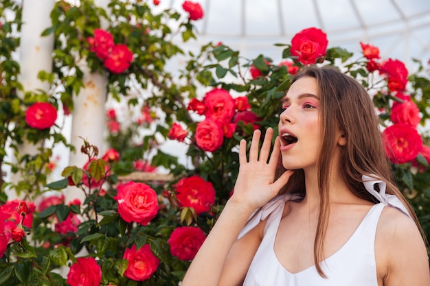 Retrato de mulher jovem e bonita chamando por alguém com a mão na boca sobre flores