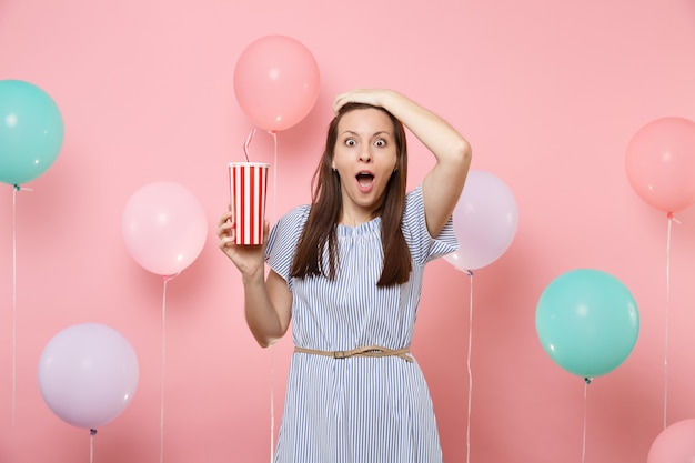 Retrato de mulher jovem e bonita chocada com vestido azul agarrado à cabeça, segurando o copo plástico de coca-cola ou refrigerante em fundo rosa pastel com balões de ar coloridos. Conceito de festa de aniversário.