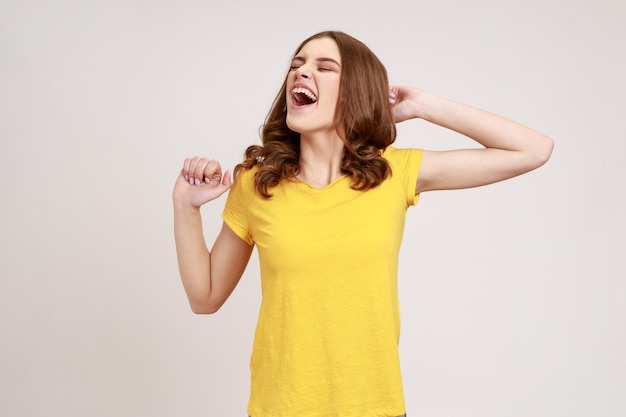 Retrato de mulher jovem e bonita com cabelos ondulados castanhos em camiseta casual amarela em pé e bocejando com os olhos fechados e braços levantados Estúdio interior isolado em fundo cinza