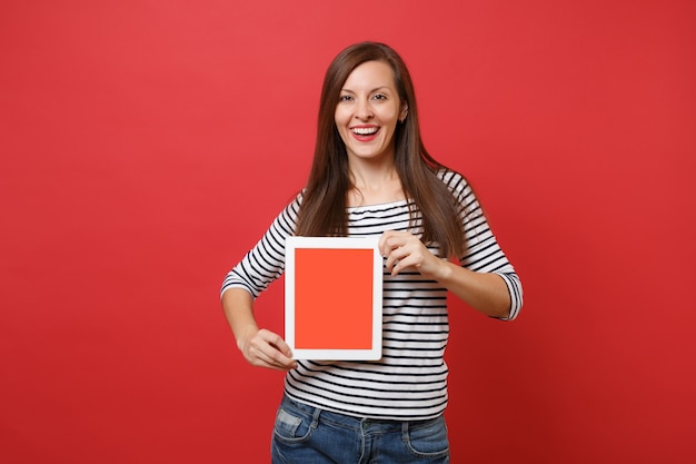 Foto retrato de mulher jovem e bonita com roupas listradas, segurando o computador tablet pc com tela vazia preta em branco isolada sobre fundo vermelho. conceito de estilo de vida de emoções sinceras de pessoas. simule o espaço da cópia.