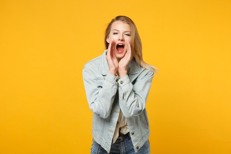 Foto retrato de mulher jovem e bonita em roupas casuais jeans, gritando com gesto de mão perto da boca isolada no fundo da parede laranja amarela no estúdio. conceito de estilo de vida de pessoas. mock up espaço de cópia.