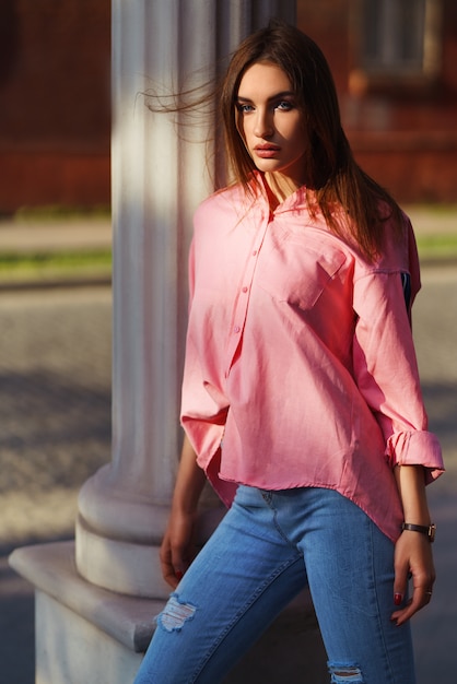 Retrato de mulher jovem e bonita em roupas casuais na rua. vestida com uma camisa rosa e calça jeans. conceito primavera / verão. hora de relaxar. garota de olhos azuis. Aproveite o pôr do sol da rua