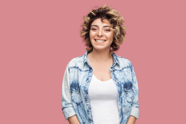 Retrato de mulher jovem e bonita feliz com maquiagem e penteado encaracolado em casual camisa azul em pé e olhando para a câmera com um sorriso. tiro de estúdio interno, isolado no fundo rosa.