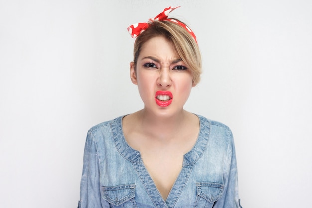 Retrato de mulher jovem e bonita insatisfeita em camisa jeans azul casual com maquiagem e bandana vermelha em pé e olhando para a câmera cerrando os dentes. tiro do estúdio interno, isolado no fundo branco.