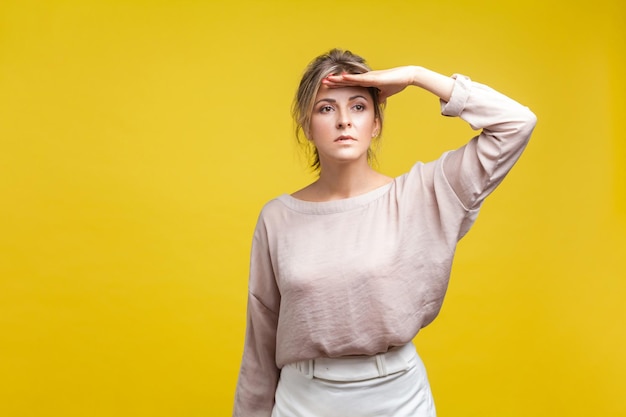 Retrato de mulher jovem e bonita observadora com cabelo louro na blusa bege casual em pé com a mão sobre a cabeça olhando para longe com olhar atento tiro de estúdio interior isolado em fundo amarelo