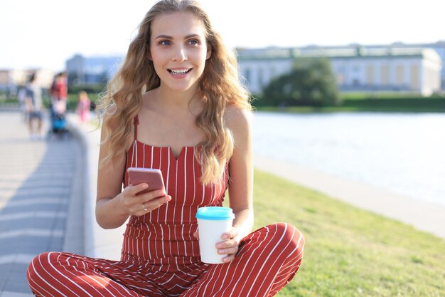 Retrato de mulher jovem e bonita sentada na margem do rio com as pernas cruzadas durante o dia de verão, usando o smartphone.