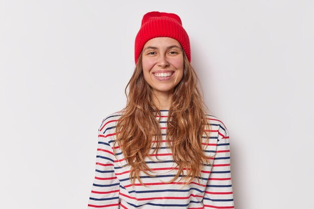 Retrato de mulher jovem e bonita sorri feliz parece ter uma conversa amigável com amigo usa jumper listrado casual de chapéu vermelho isolado sobre fundo branco. Conceito de emoções felizes