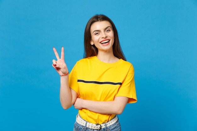 Retrato de mulher jovem e bonita sorridente em roupas casuais vívidas, olhando para a câmera, mostrando o sinal de vitória isolado no fundo da parede azul brilhante no estúdio. conceito de estilo de vida de pessoas. mock up espaço de cópia.