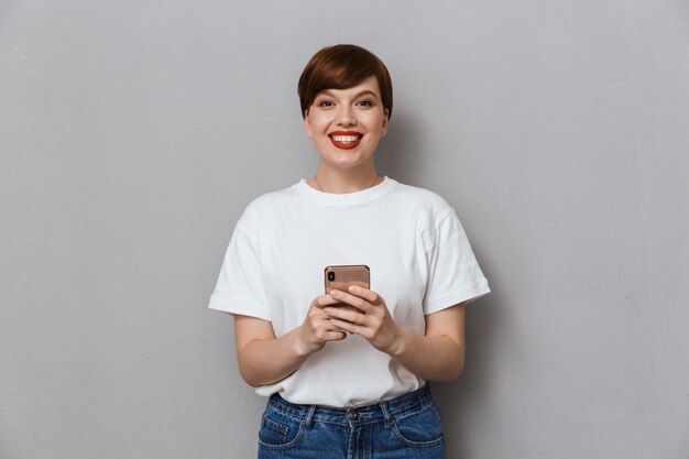 Retrato de mulher jovem e bonita sorrindo e segurando o celular isolado sobre uma parede cinza