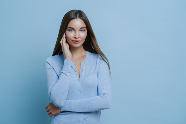 Retrato de mulher jovem e bonita toca o rosto suavemente, usa jumper azul, tem uma aparência atraente