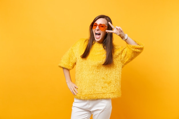 Retrato de mulher jovem e feliz animada com suéter de pele e óculos coração laranja mostrando sinal de vitória isolado em fundo amarelo brilhante. emoções sinceras de pessoas, conceito de estilo de vida. área de publicidade.