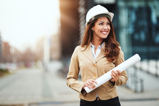 Foto retrato de mulher jovem engenheiro com capacete branco e plantas nas mãos no canteiro de obras.