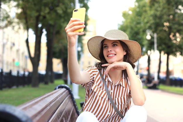 Retrato de mulher jovem, fazendo selfie pelo telefone, sentado no banco na rua da cidade.