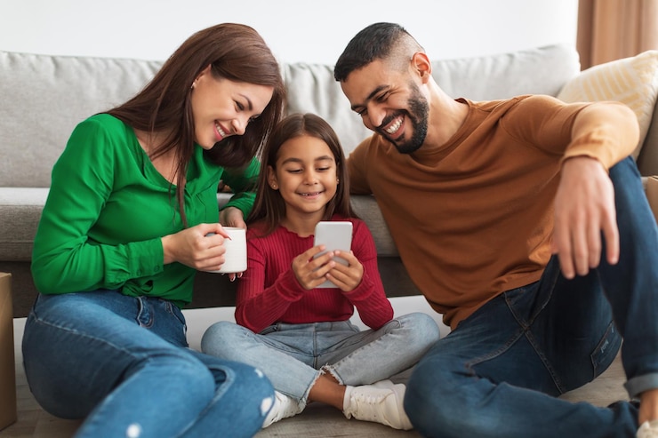 Foto retrato de mulher jovem feliz, homem e menina segurando e usando smartphone sentado no chão na sala de estar. pessoas passando um tempo juntas, assistindo a vídeos, escolhendo presentes, compartilhando gadgets com filhas