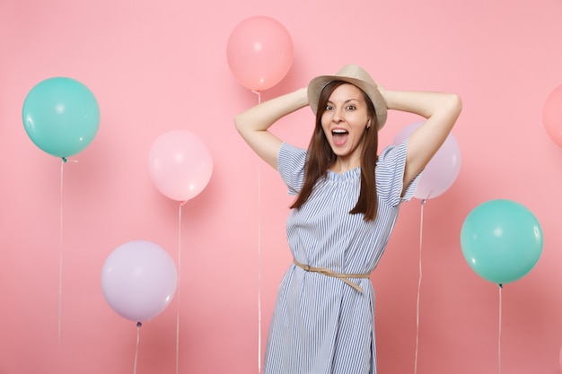 Retrato de mulher jovem feliz radiante com chapéu de palha de verão e vestido azul, mantendo as mãos perto da cabeça no fundo rosa com balões de ar coloridos. Emoções sinceras de pessoas de festa de férias de aniversário.