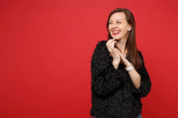 Retrato de mulher jovem muito alegre sorridente em suéter de pele preta olhando de lado isolado no fundo da parede vermelha brilhante no estúdio. Emoções sinceras de pessoas, conceito de estilo de vida. Simule o espaço da cópia.