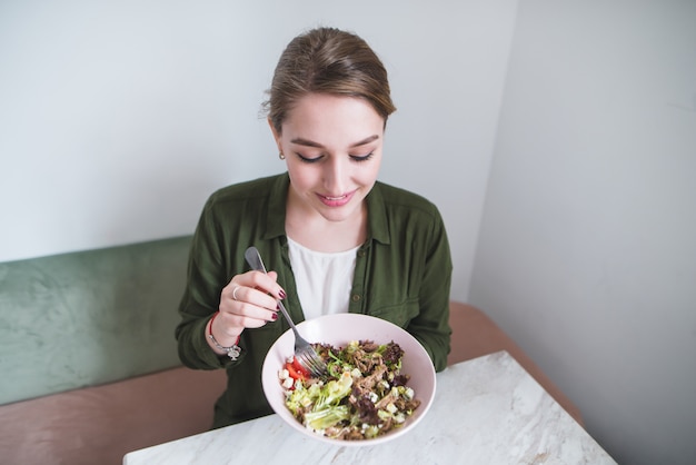 Retrato de mulher jovem positiva com prato de salada nas mãos no interior brilhante. Alimentação saudável
