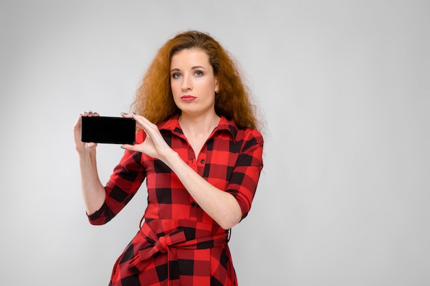 Retrato de mulher jovem séria ruiva linda no vestido xadrez, mostrando o telefone móvel em fundo cinza