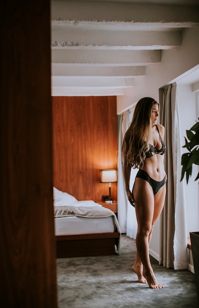 Retrato de mulher jovem sexy em lingerie em pé pela janela no quarto