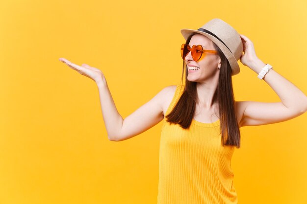 Retrato de mulher jovem sorridente com chapéu de palha de verão, óculos laranja apontando para a mão direita de lado copie o espaço isolado em fundo amarelo. Emoções sinceras de pessoas, conceito de estilo de vida. Área de publicidade.